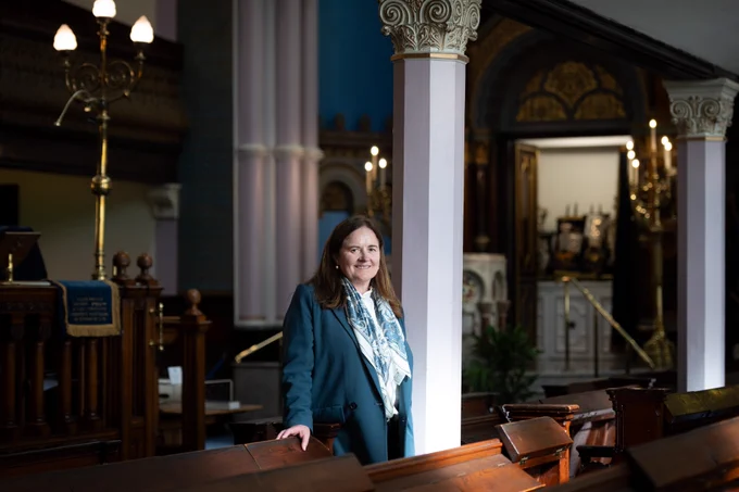 A person in a teal colour jacket standing next to a white supporting column. The photo is from Garnethill Synagogue in Glasgow