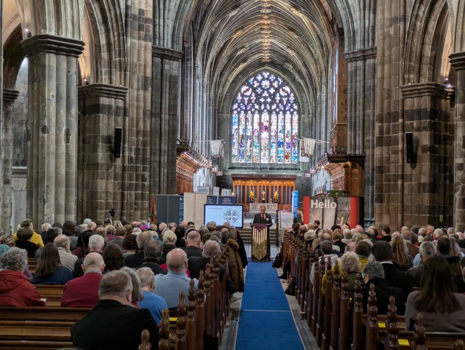 Interior of a churchwith pews full of people. at the back is a large stained glass window. This is Paisley Abbey