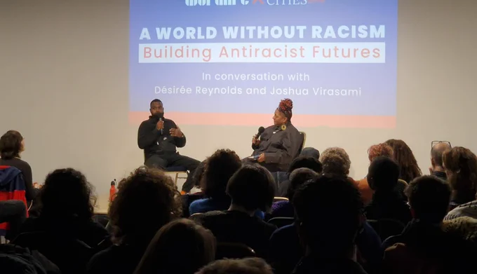 A photo of two people in conversation at a public event. Both are sat down and one is holding a microphone. In the background is a projection with the words "A WORLD WITHOUT RACISM. Building Antiracist Futures"