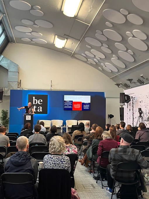 A photo of a conference of people listening to a speaker on stage. There are a number of people facing the stage as one person talks at a black podium. There are four white chairs and a white table on the stage and behind the chairs is a tv screen.