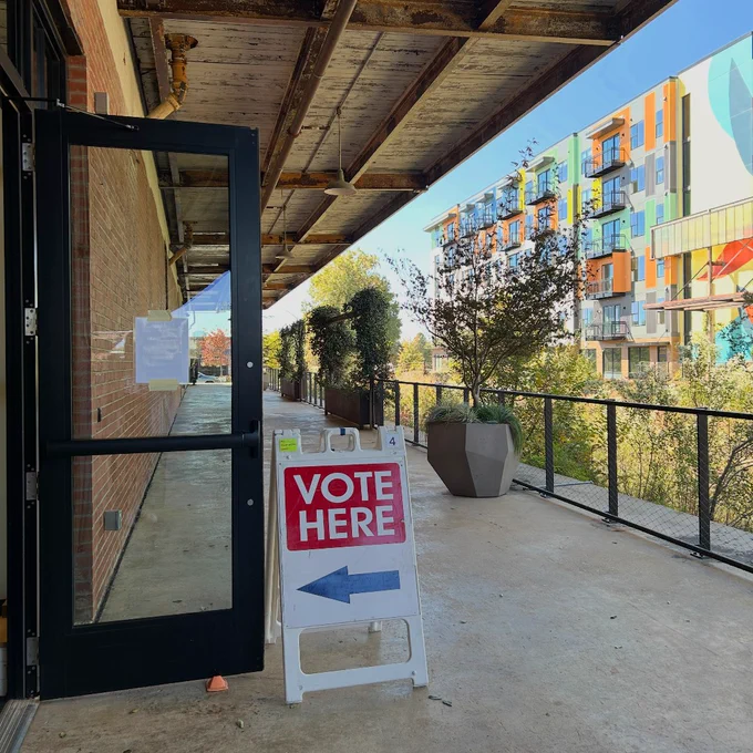 A "Vote Here" sign is placed outside an open door along a covered walkway, with modern apartment buildings visible in the background.