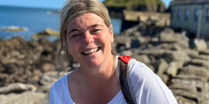 A colour image of a smiling person with tied back blonde hair and wearing a white top, with a red and black bag handle visible on one shoulder. There are rocks and water visible in the background.