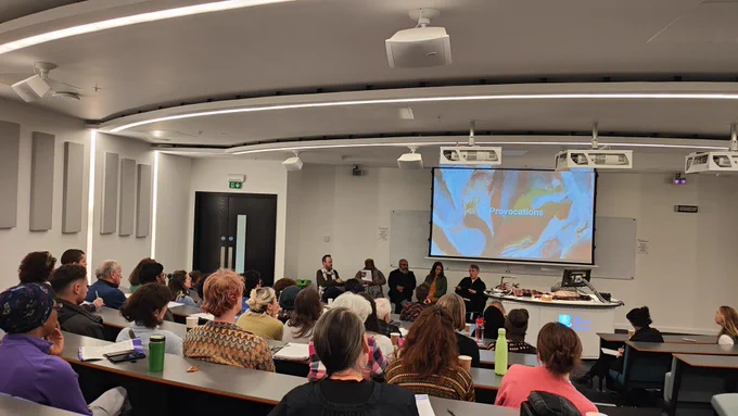 A photo of a group of people in a lecture theatre watching a panel discussion with five people sitting at the front