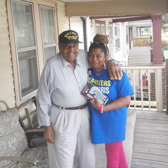 A picture of two people in Michigan who are voting for Kamala Harris. The man wears a World War II veteran hat. The woman is wearing a Teamsters for Harris shirt.