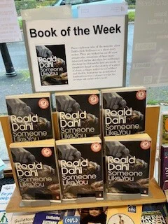 Image is a photograph of Horsley Library's Book of the Week display, a plastic stand featuring 6 copies of Roald Dahl's "Someone Like You". Above the display is a laminated advertisement in landscape format. Said advertisement is titled: 'Book of the Week' and has a picture of the Book's cover on the left and the text of the blurb, reading: 'These eighteen tales of the macabre show Dahl's dark brilliance as a short story writer.  They are wicked (as an old man attracts the attentions of those more interested in his skin than his wellbeing), shocking (as distasteful bets are made - a daughter's hand on the identity of a glass of claret, a finger risked for a Cadillac) and blackly humorous (as a cuckolded husband receives a chance to take his revenge out on his wife's neck).'

