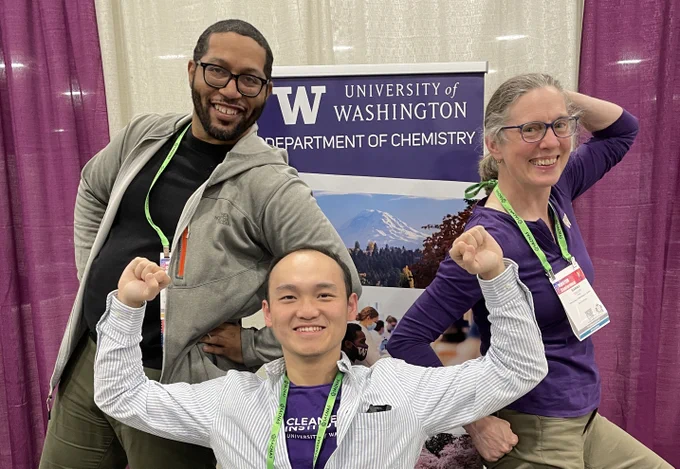 The image is a photograph of three smiling people in front of a purple sign that says “University of Washington, Department of Chemistry”.