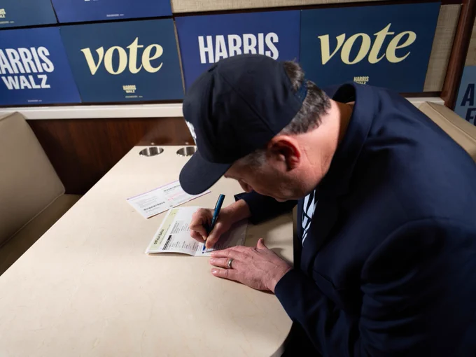 Second Gentleman Doug Emhoff marking his ballot to vote for Vice President Kamala Harris and Governor Tim Walz with signs reading "Vote" and "Harris Walz" in the background. He is wearing a dark blue Harris-Walz cap and shirt under his dark blue blazer. 