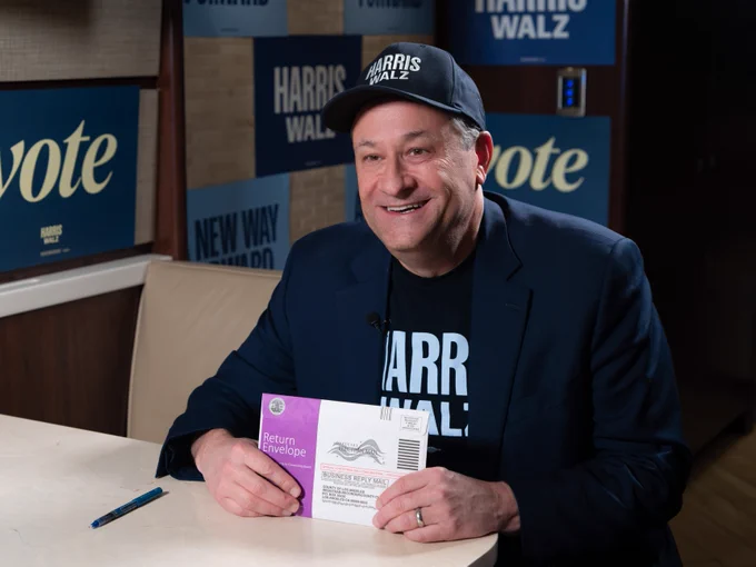 Second Gentleman Doug Emhoff smiling while holding up his ballot return envelope to vote for Vice President Kamala Harris and Governor Tim Walz. There are signs reading "Vote" and "Harris Walz" in the background. He is wearing a dark blue Harris-Walz cap and shirt under his dark blue blazer. 