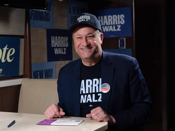 Second Gentleman Doug Emhoff smiling with an "I Voted" sticker attached to the lapel of his blazer. There are signs reading "Vote" and "Harris Walz" in the background. He is wearing a dark blue Harris-Walz cap and shirt under his dark blue blazer. 