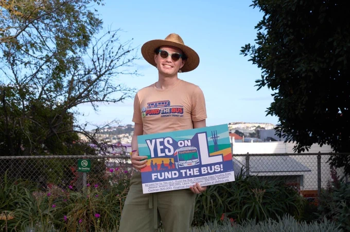 Aidan in a Fund the Bus t-shirt, sunglasses and sun hat holding a "Yes on L, Fund the Bus" window sign in a garden with a San Francisco landscape in the background