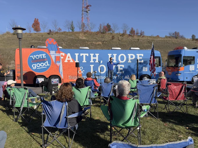 Chris Deluzio speaking into a handheld microphone in front of an orange and blue tour bus. A group of around 10 people are sitting and watching.
