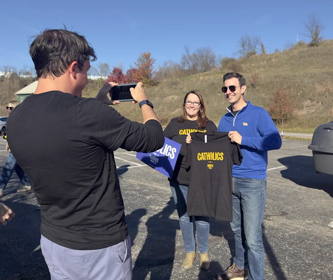 Chris Deluzio poses with a woman for a photo that a man is taking on a phone. Deluzio is holding a black t-shirt with the text “Catholics for Harris.”