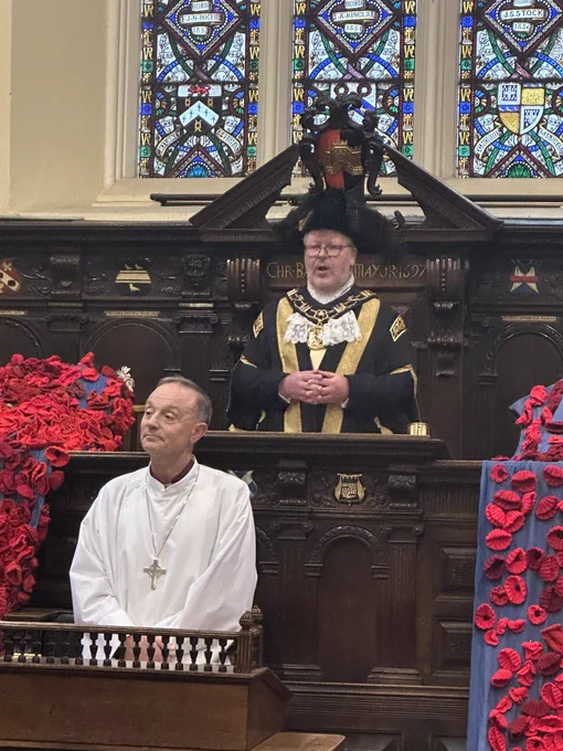 The Lord Mayor in black and gold@robes stands behind the bishop in a white lab with displays of poppies either wide of them