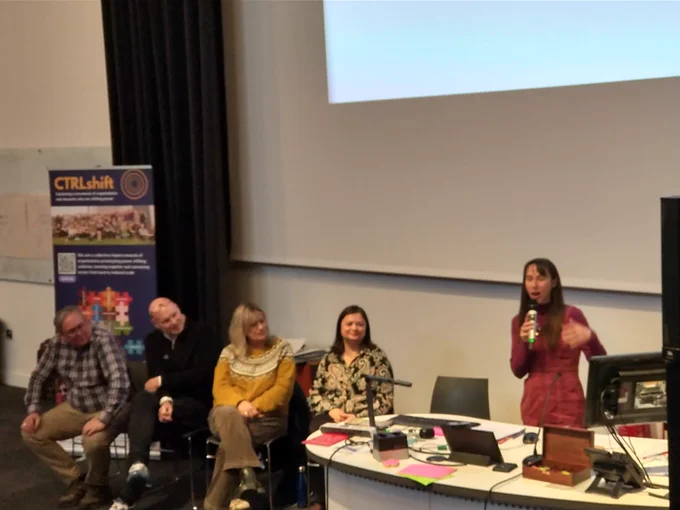 A photo of five people speaking at the front of a lecture theatre. Four are sat down, and one is stood at a lectern with a microphone.