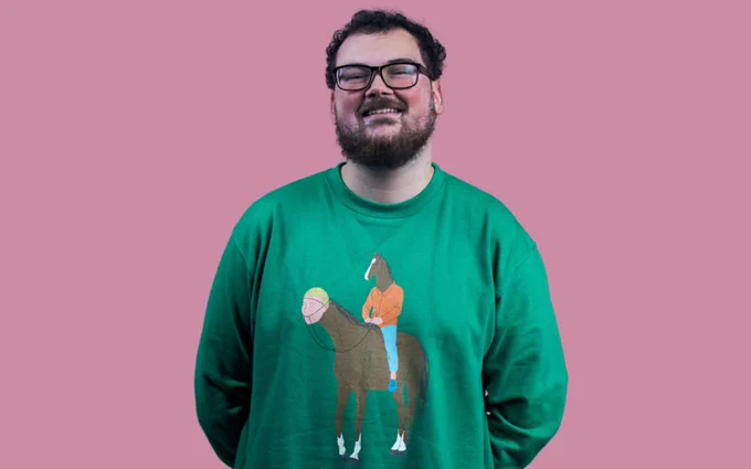 Alex Mitchell smiling, wearing glasses in a green Bojack Horseman sweatshirt where a man with a horse's head is riding a horse with a human head. Alex is stood against a pink background.