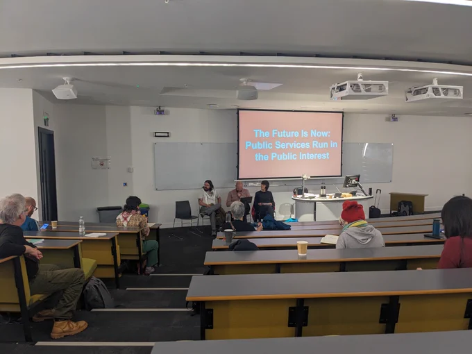 A photo from the back of a lecture theatre, with three people sat at the front with microphones. On a screen behind them are the words "The Future Is Now: Public Services Run in the Public Interest"