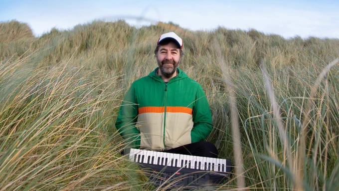 David O'Doherty stood in a long grass wearing a green and beige jacket with a keyboard.