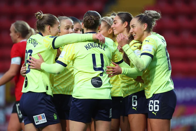 Southampton FC Women's players huddle in celebration after Tara Bourne's winning goal against Charlton at The Valley, all wearing the yellow away kit with navy shorts