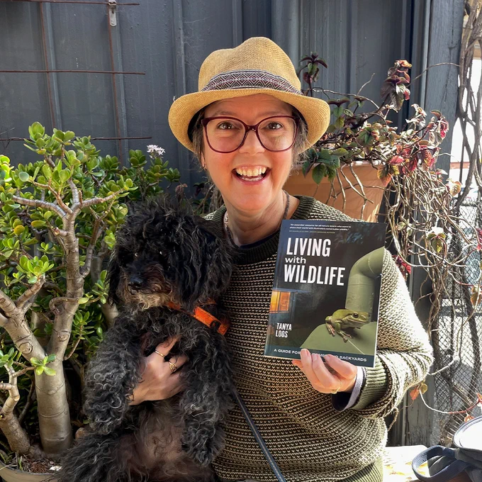 Tanya Loos sitting with her dog Valli on a bench, smiling and holding a copy of her book Living with Wildlife.