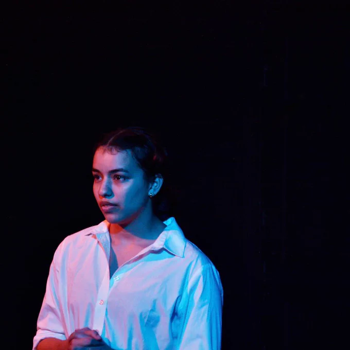 A dress rehearsal photo of a play. A femme presenting person with dark hair and eyes is wearing a white shirt. Blue and pink light bounces off of their shirt and face. They are expressionless.