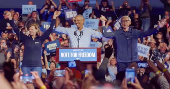 Tammy Baldwin, Barack Obama, and Tony Evers all smile while raising their hands together. Behind them, a huge crowd cheers.