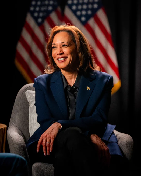 Vice President Kamala Harris seated and smiling, with two American flags in the background.