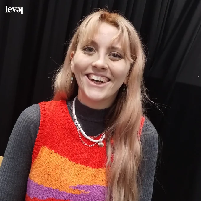 A person smiles broadly at the camera. They have long blonde hair. They wear earing and a nose ring. The have a grey long sleeved top on with a colourful tank top over it, in red, orange and purple. They wear several necklaces over this. The background is a black curtain.