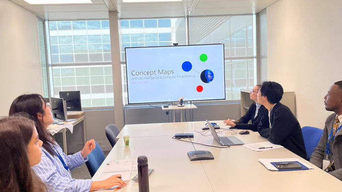 Five professionals sitting at an office desk watching a screen where a presentation is being shown; the presentation’s title "Concept Maps" and an image with circular graphs are visible on screen. An open laptop, water bottles, paper cups, pens and notebooks are on the desk. 