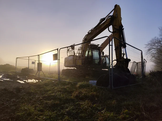 A large digging machine, ensconced in a metal-fenced playpen, is silhouetted by the morning sun. The surround is a misty field, fading away into the background.