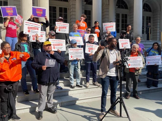 The Public Advocate speaks in front of a rally on the steps of City Hall, raising one hand in emphasis. Advocates hold signs with messages like "Vote NO on proposals 2-6. Protect New York City democracy. #Nix2Through6"