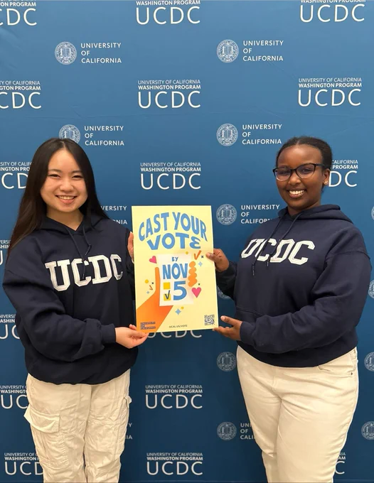 From left to right: UC Merced student Isabella and UC Berkeley student Idyl, both participants in the University of California Washington program, stand in front of a UCDC branded backdrop wearing UCDC hoodies and holding a yellow poster with blue text that says, “Cast your vote by Nov. 5.” 
