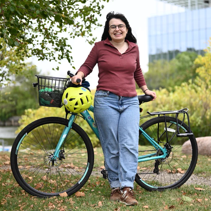 A young woman stands smiling with her bike in a park overlooking part of the river. The woman is wearing a red long sleeved shirt and blue jeans with brown sneakers, and has light skin, long dark brown hair, and circular glasses. Her bike is a blueish green Reid hybrid bike with disk brakes, a black metal basket on the front handlebars, and a cargo rack on the back., and there are some dry brown leaves on the grassy ground by the bike.