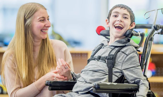 A young smiling wheelchair user is in his Matrix-style chair. A smiling woman caregiver sits beside him.