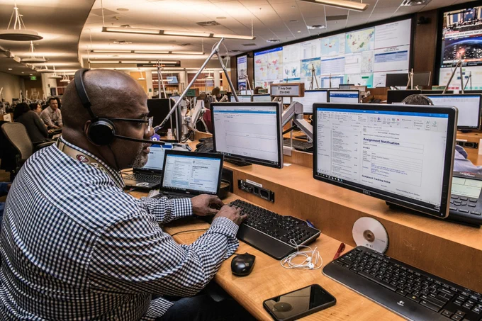 A man sits in front of a computer in a room filled with screens, and people. The room looks like a monitoring facility for IT or incident response. The man has a checkered shirt and a headset. Photo by CDC on Unsplash
