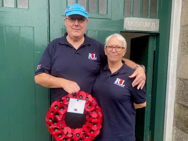 Two people stand in front of green doorway holding a poppy wreath.