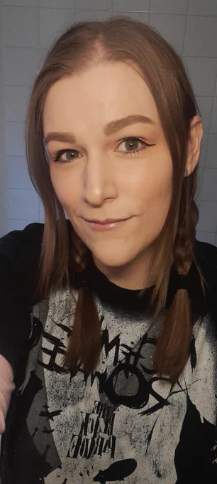 A selfie of Lav facing the camera. Her head, shoulders, and most of her t-shirt are in the shot. Behind her are white bathroom tiles. Her hair is brown and in braids, with strands on either side hanging loose at the front. Lav is wearing makeup, with winged black eyeliner and pink lipstick. The t-shirt she is wearing is black and white, and the text on it reads "My Chemical Romance The Black Parade."