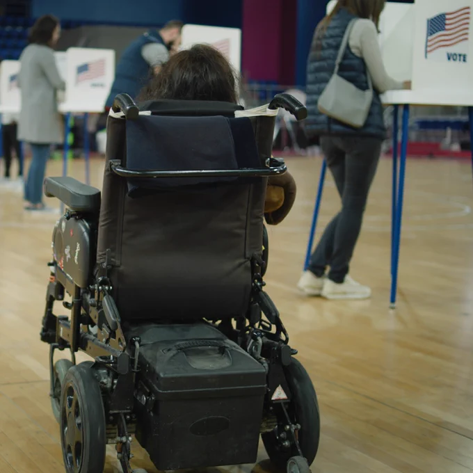 A woman in a power wheelchair enters a voting facility. To her right are people standing to vote at tables.