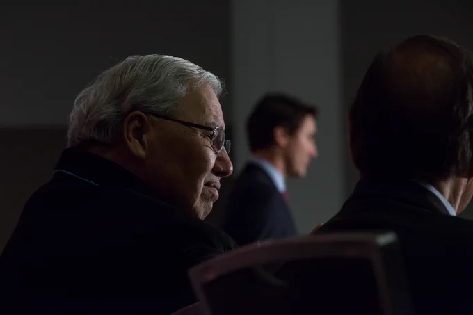 The Honourable Murray Sinclair is seated next to a person, at an event in Ottawa, Ontario. The side of his face is in focus, and Prime Minister Justin Trudeau is standing in the background.