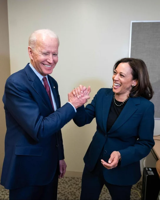 President Joe Biden and Vice President Kamala Harris, both wearing suits, smile and clasp hands.