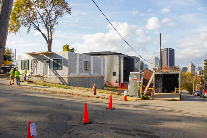 Construction workers stand near a ladder in front of the first level of a modular home. The boxes were built off site and placed on a concrete pad at the site. There are traffic cones preventing people from going down the street, and a view of downtown Pittsburgh is seen in the background.
