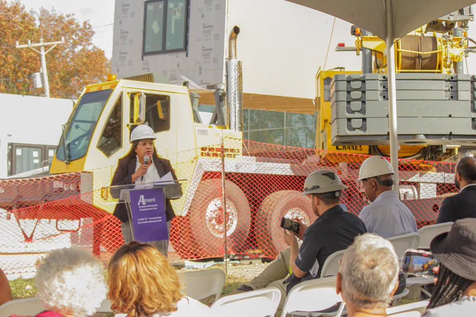 Quianna Wasler, Chief Housing Officer of the URA, talks to a group of people. She is wearing a hard hat and stands in front of a construction site where part of a modular home is being placed on a concrete pad. 