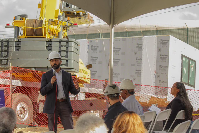 Kyle Chintalapalli, Chair of the URA Board of Directors, talks to a group of people. He is wearing a hard hat and stands in front of a construction site where part of a modular home is being placed on a concrete pad. 