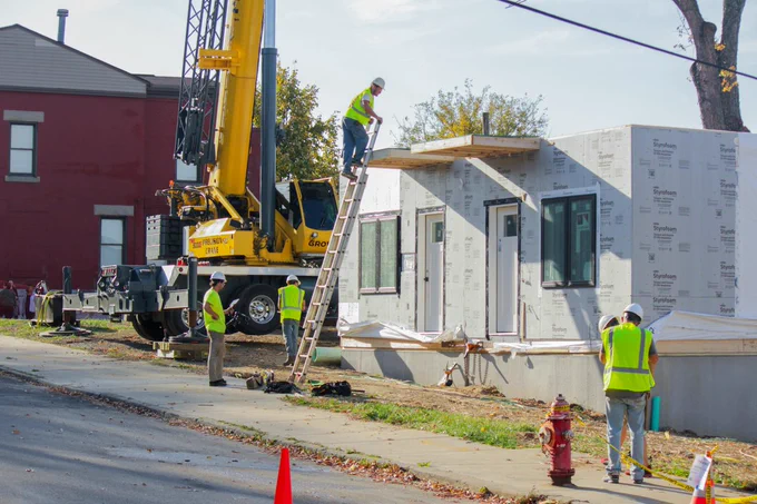 Construction workers stand around the first level of a modular home that is being placed on a concrete pad. A crane that lifts the boxes of the home is seen in the background. One construction worker is at the top of a ladder, spotted by another at the bottom.