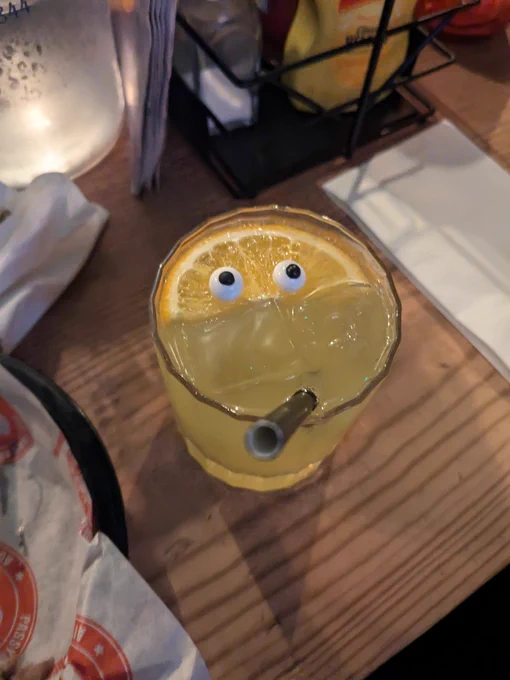 An orange cocktail with a half slice of orange laid flat on top, the orange has 2 googly eyes on it making the whole cocktail look like a face