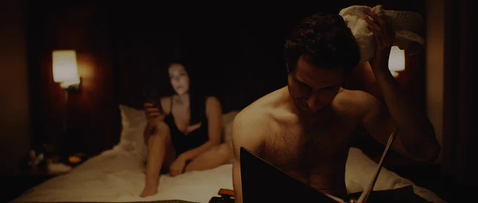 Image is set in a bedroom with a darker mood. A shirtless man can be seen in the front of the photo sitting on the end of the bed holding an ice pack on his head and studying a binder. In the background is a woman wearing pjs and on her phone. Photo by Carolina Bayon.