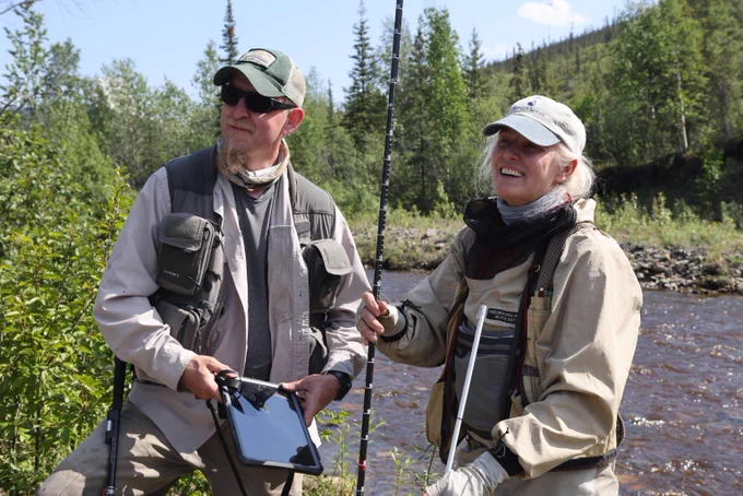 Two people stand with equipment in front of a river and trees