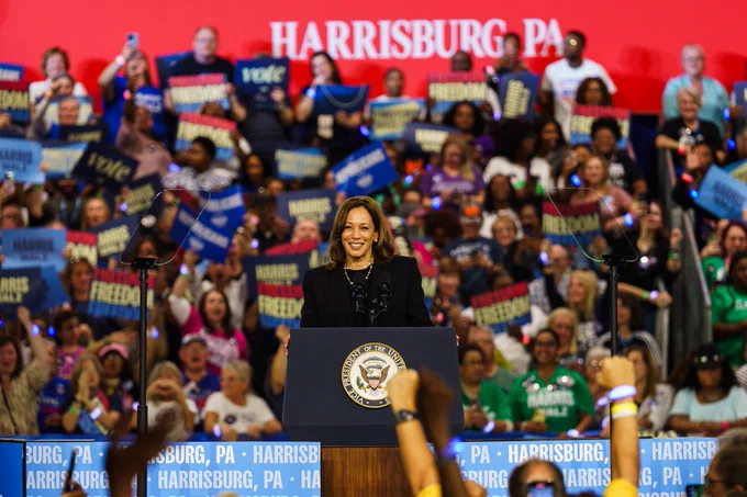 Vice President Kamala Harris standing on stage behind a podium at a campaign event in Harrisburg, Pennsylvania. In the background the audience is holding up signs and clapping. 