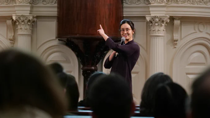 A presenter at 2023 StreetTalk 10-in-1 points up and to her right with her left hand, while holding a microphone in her right hand. She is smiling at the audience and holding the microphone up to her mouth.