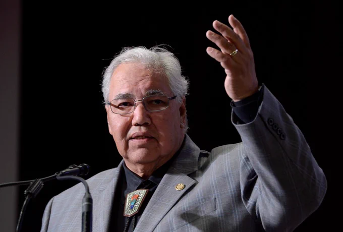 A close- up of Murray Sinclair speaking at a podium. Several microphones are in front of him and he is raising his left, outstretched hand.