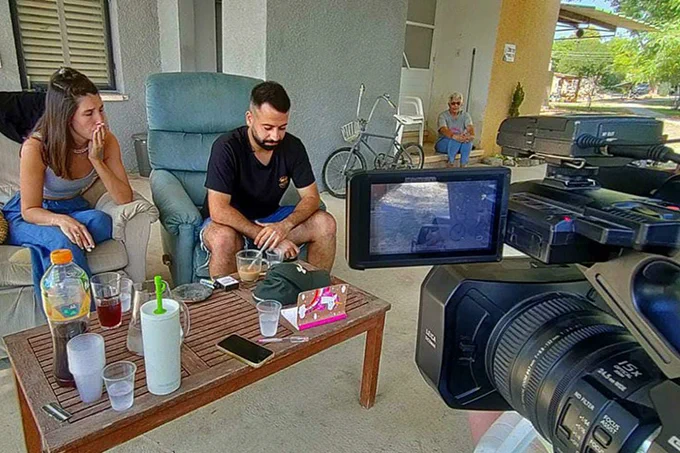 A woman and man sit on a couch with a large TV camera trained on them.